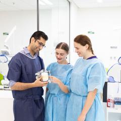3 Dental students in a lab wearing blue scrubs and discussing a device